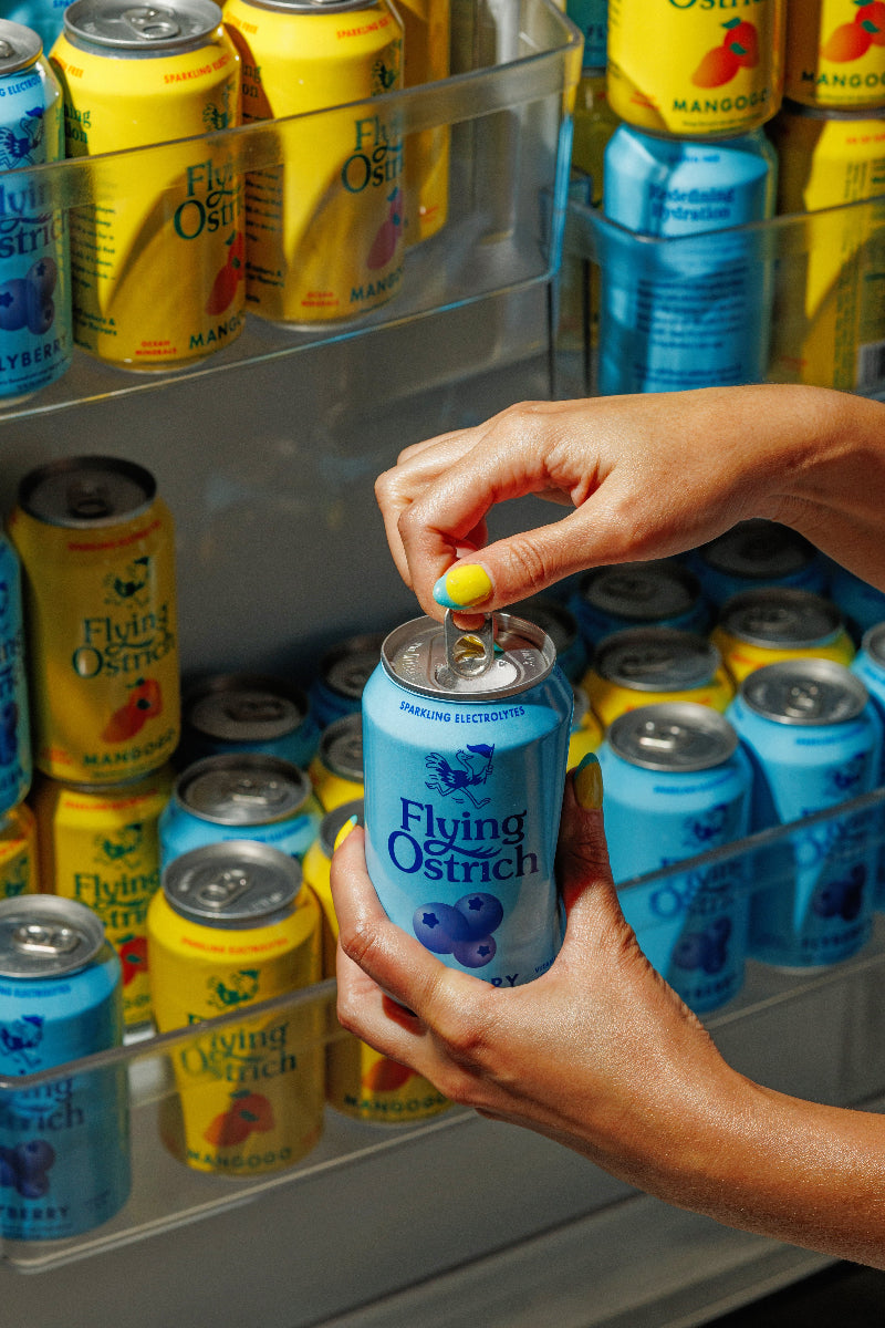Hand holding a blue can of Flyberry in front of a refrigerator stocked with various cans.